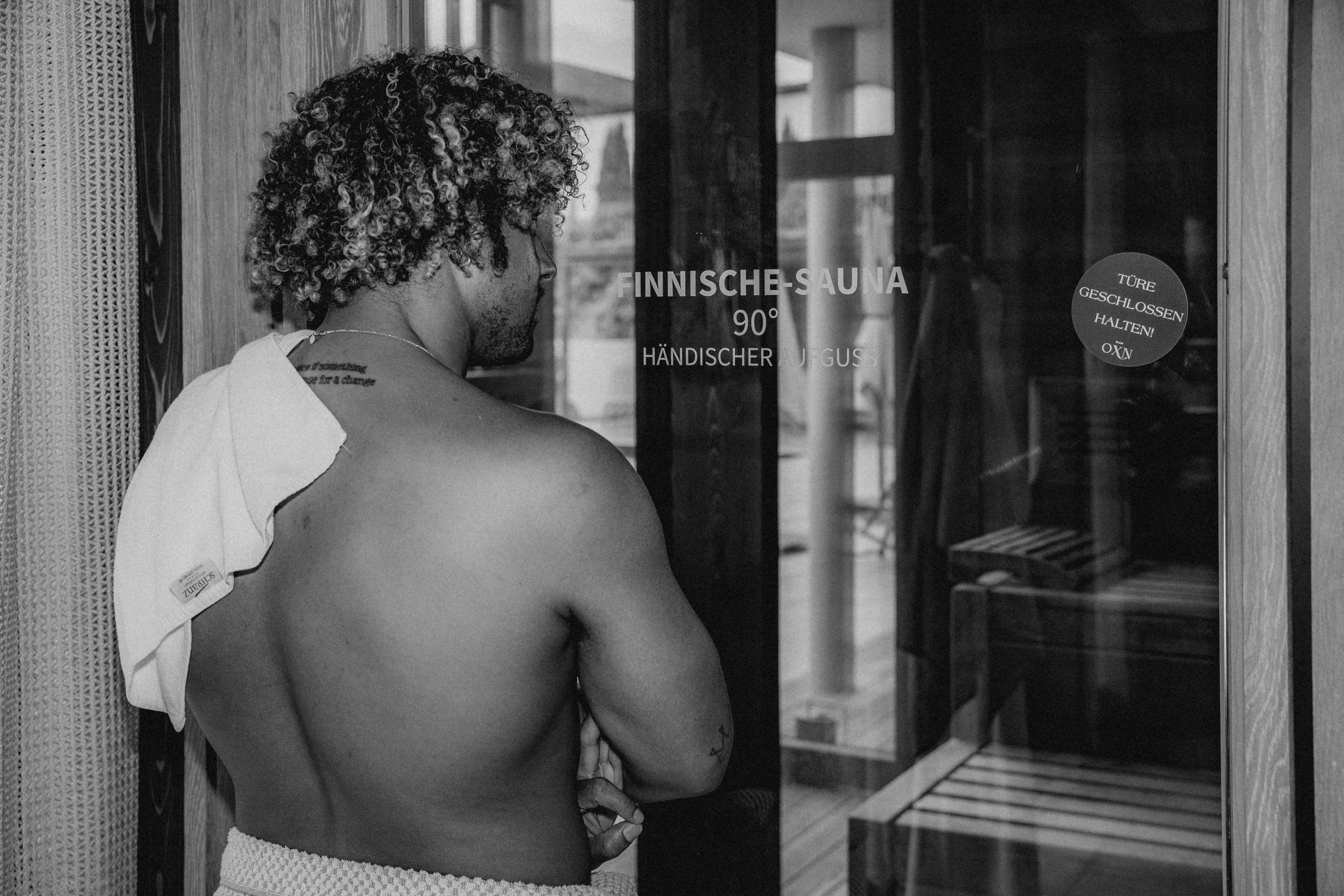 A person with curly hair stands near a doorway, looking contemplatively at something outside the frame.