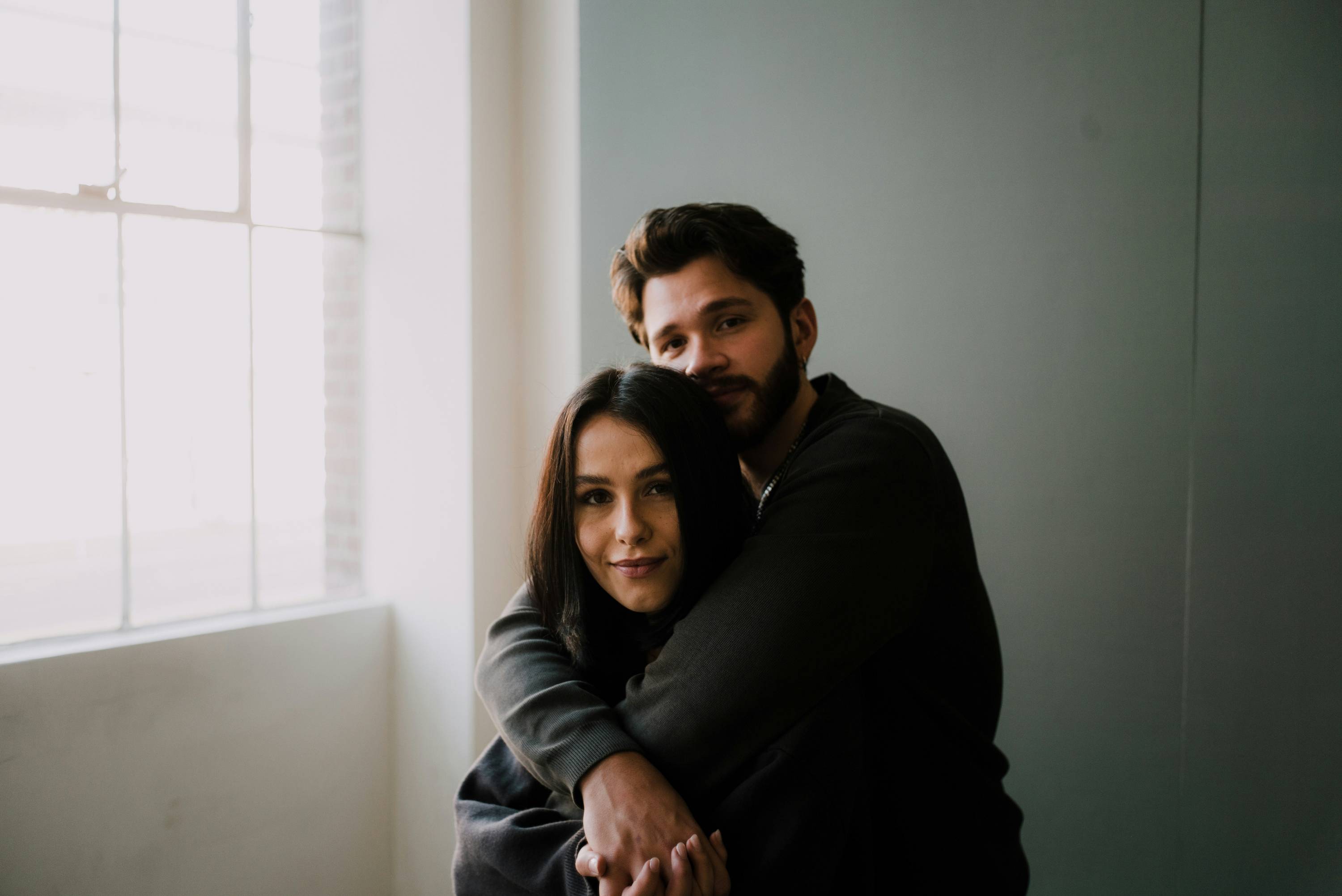 A Couple in a bright lit studio looks a the viewer while in a loose embrace