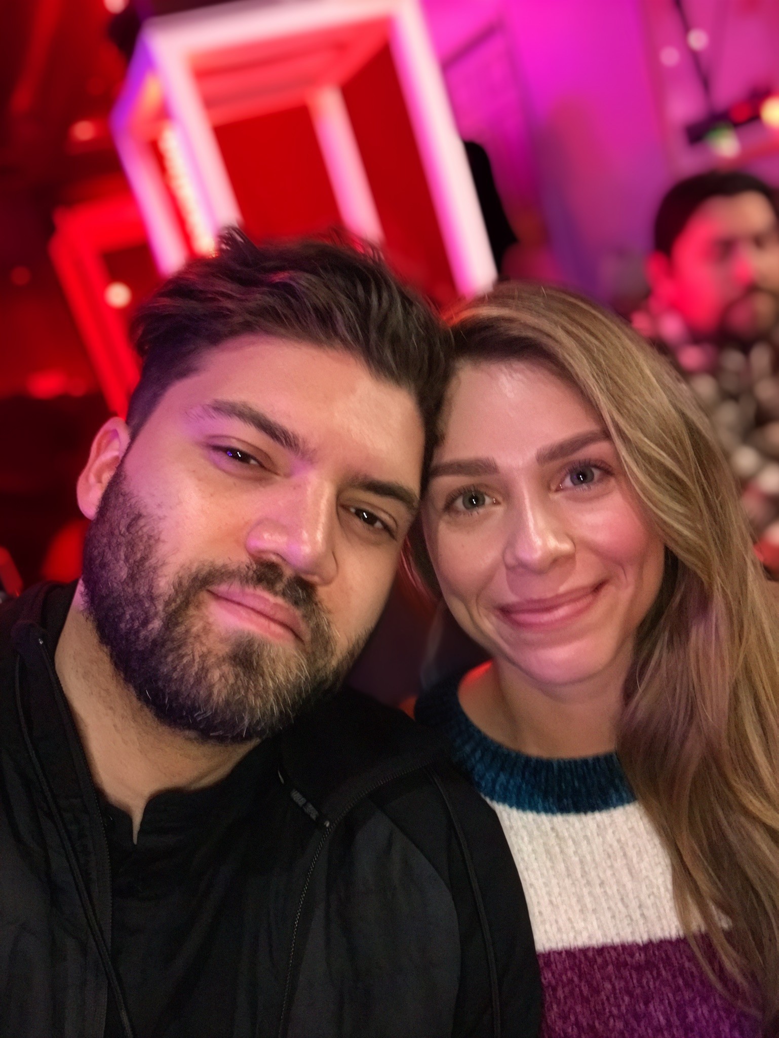 A groom in a dark suit carries a bride in a white dress across a sunny, grassy field. The couple shares a joyful, intimate moment with trees in the background.