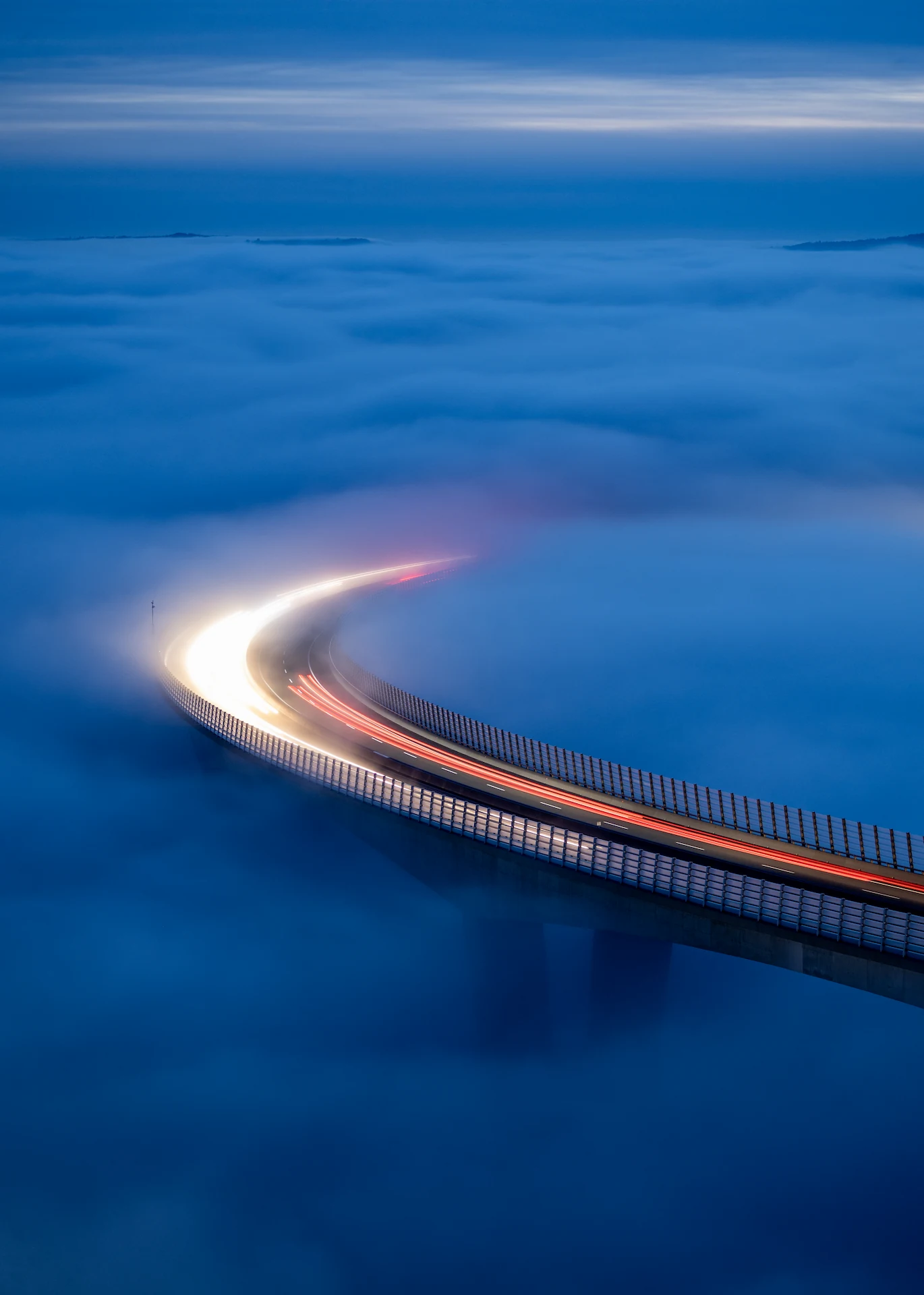 A long-exposure shot of the Črni Kal viaduct in Slovenia at twilight, showing light trails from vehicles on the curved bridge as it cuts through a thick, blue layer of fog.