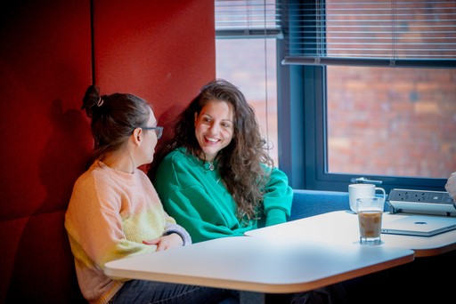 Two people sit together in a booth beside a window, talking at a table with coffees, a laptop and a notebook in a relaxed workspace with warm lighting.