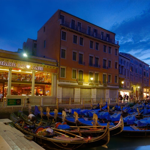 威尼斯傍晚时分的运河景致，贡多拉停泊在岸边，旁边是灯火通明的 Hard Rock Cafe 和色彩缤纷的建筑。