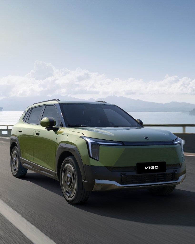 A green SUV driving on a scenic road with mountains and a cloudy sky in the background.
