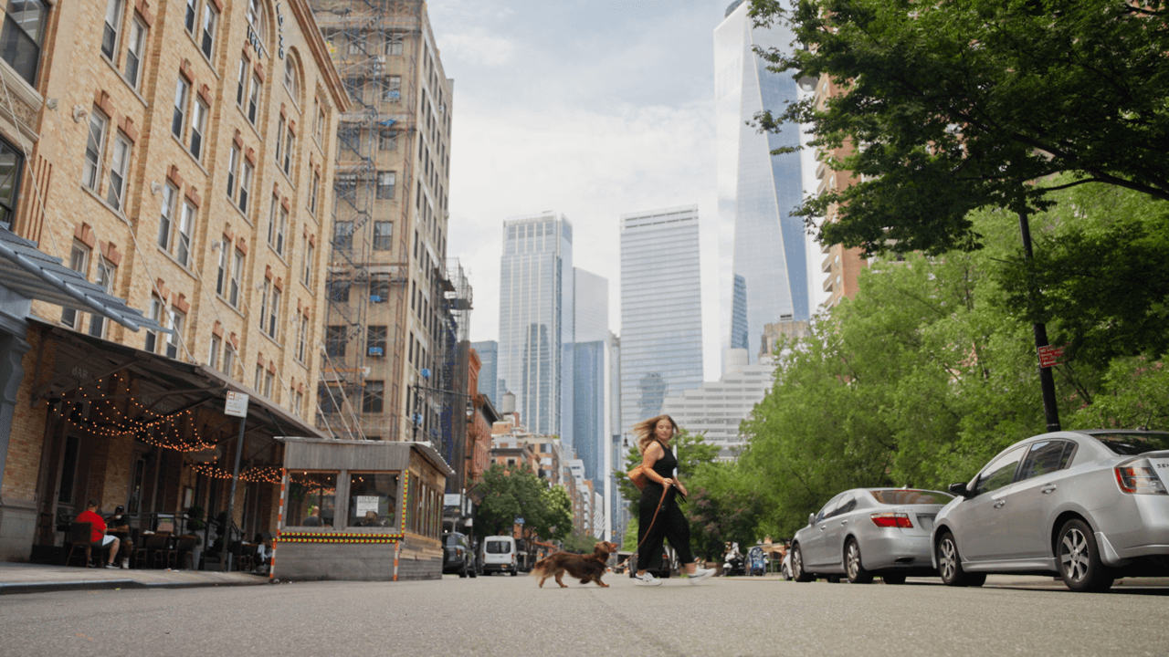 Urban street scene with tall buildings, trees, and people walking on the sidewalk