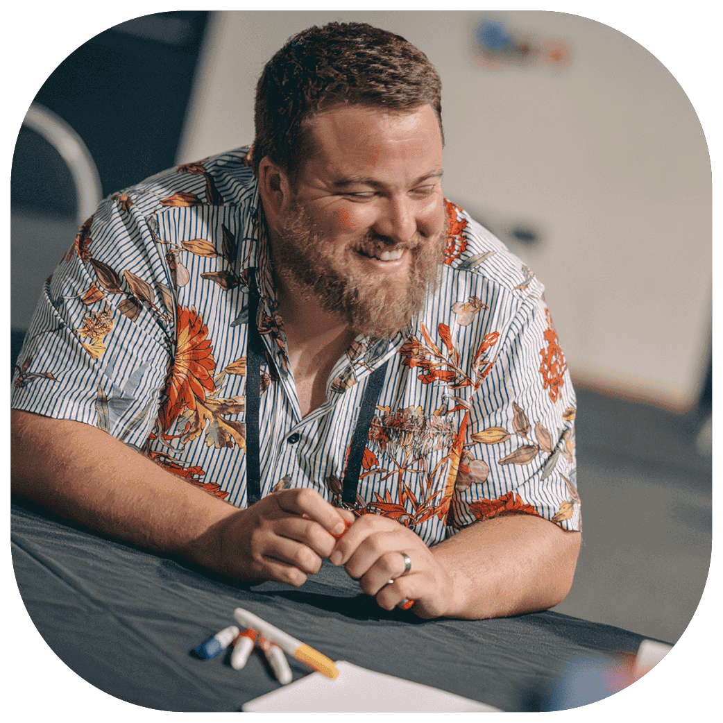 Person standing and gesturing toward a wall of sticky notes while holding a marker, wearing a lanyard in a meeting room
