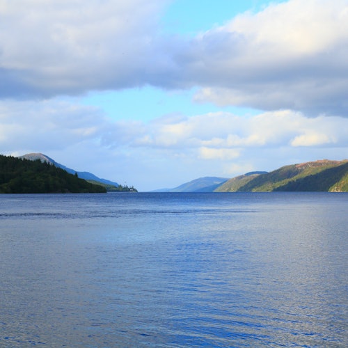 A calm lake surrounded by tree-covered hills under a partly cloudy sky.