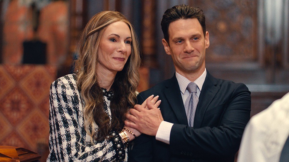 A medium shot of a woman and man standing close together in a wood-paneled room. The woman, wearing a houndstooth blazer and gold bracelets, clutches the man's arm with a strained, intense smile. The man, in a dark suit and tie, looks forward with an uncomfortable, tight-lipped grimace. They appear to be forcing polite expressions in a formal setting.