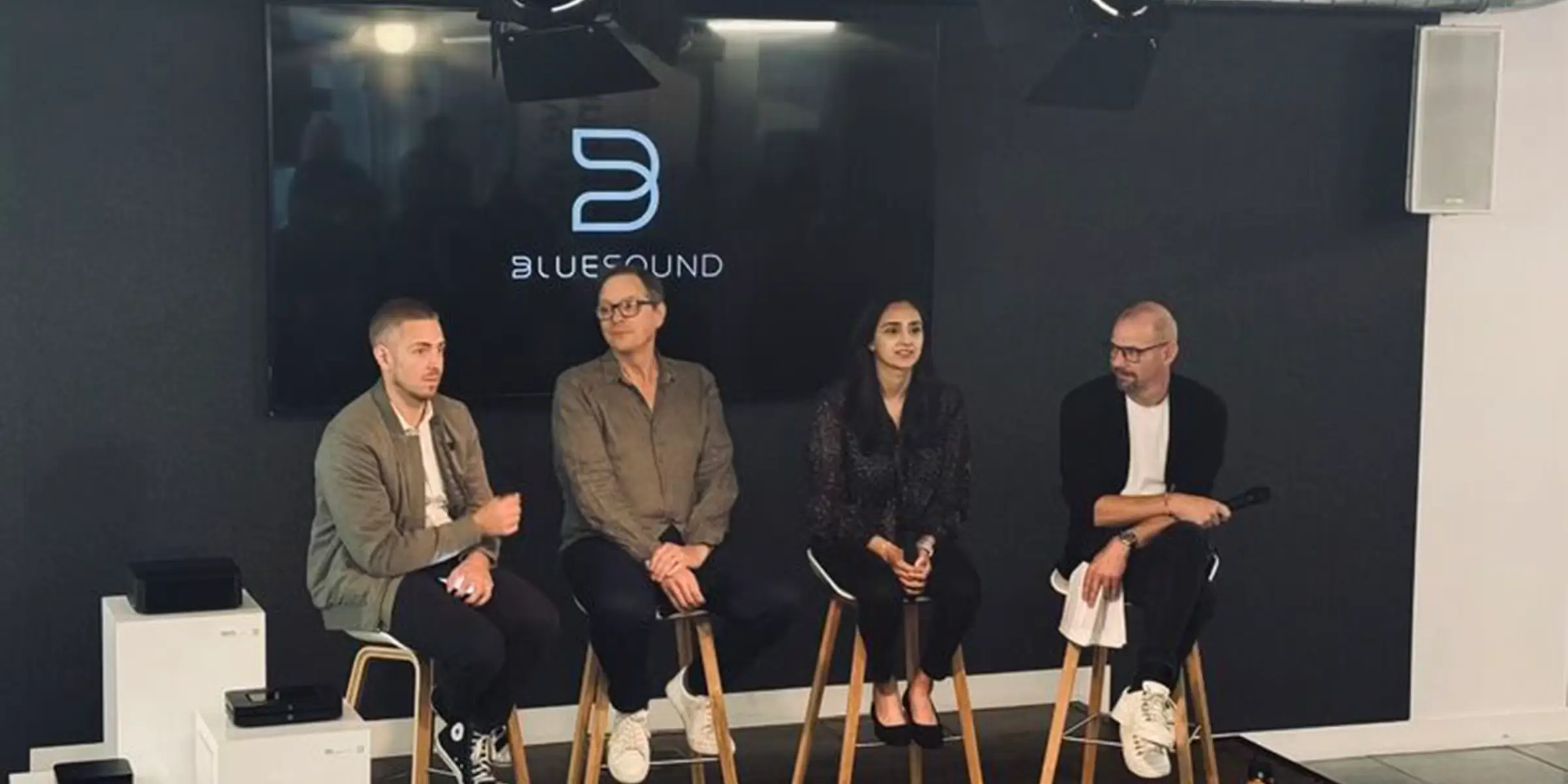 Panel discussion on a small stage with five speakers seated in front of a screen showing the Bluesound logo.