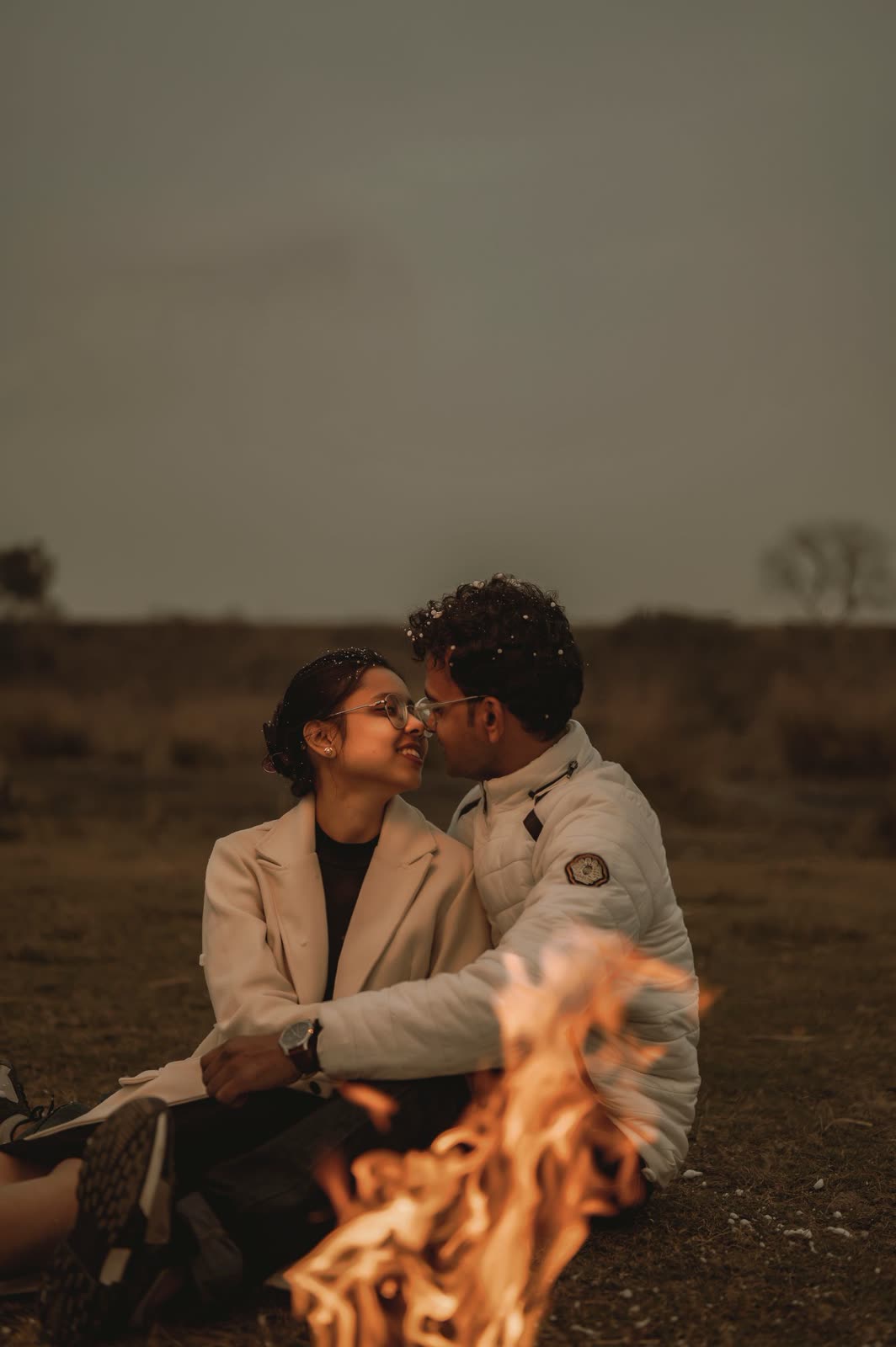 Couple in coats sitting close together beside a small fire at dusk