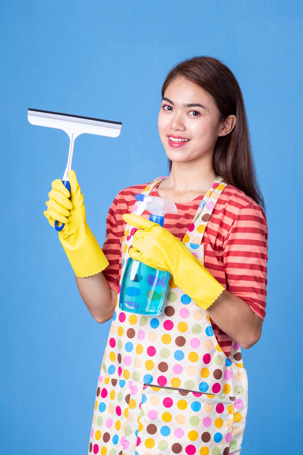 Smiling woman holding a squeegee and a spray bottle, wearing yellow gloves and a colorful apron, standing against a blue background, ready to clean windows