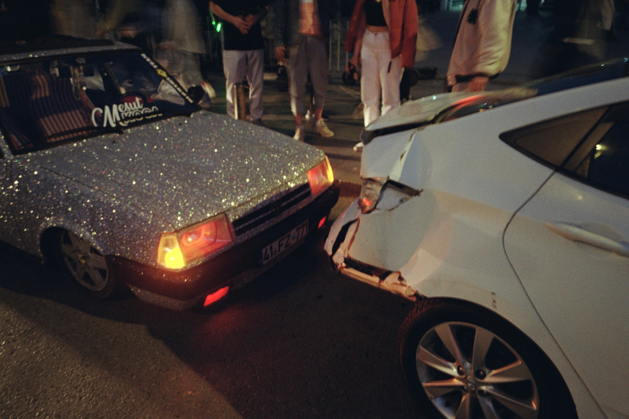 A glittery car with illuminated headlights has collided with a white sedan, resulting in noticeable rear damage, surrounded by onlookers on a nighttime street.