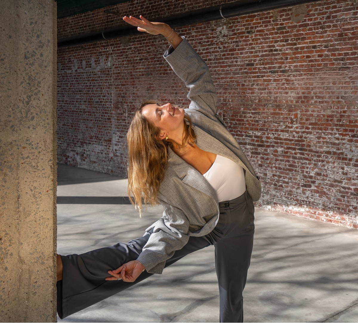 A person wearing a gray suit performs a stretching exercise against a concrete pillar in an industrial-style building with exposed brick walls, emphasizing the importance of burnout prevention through physical activity.