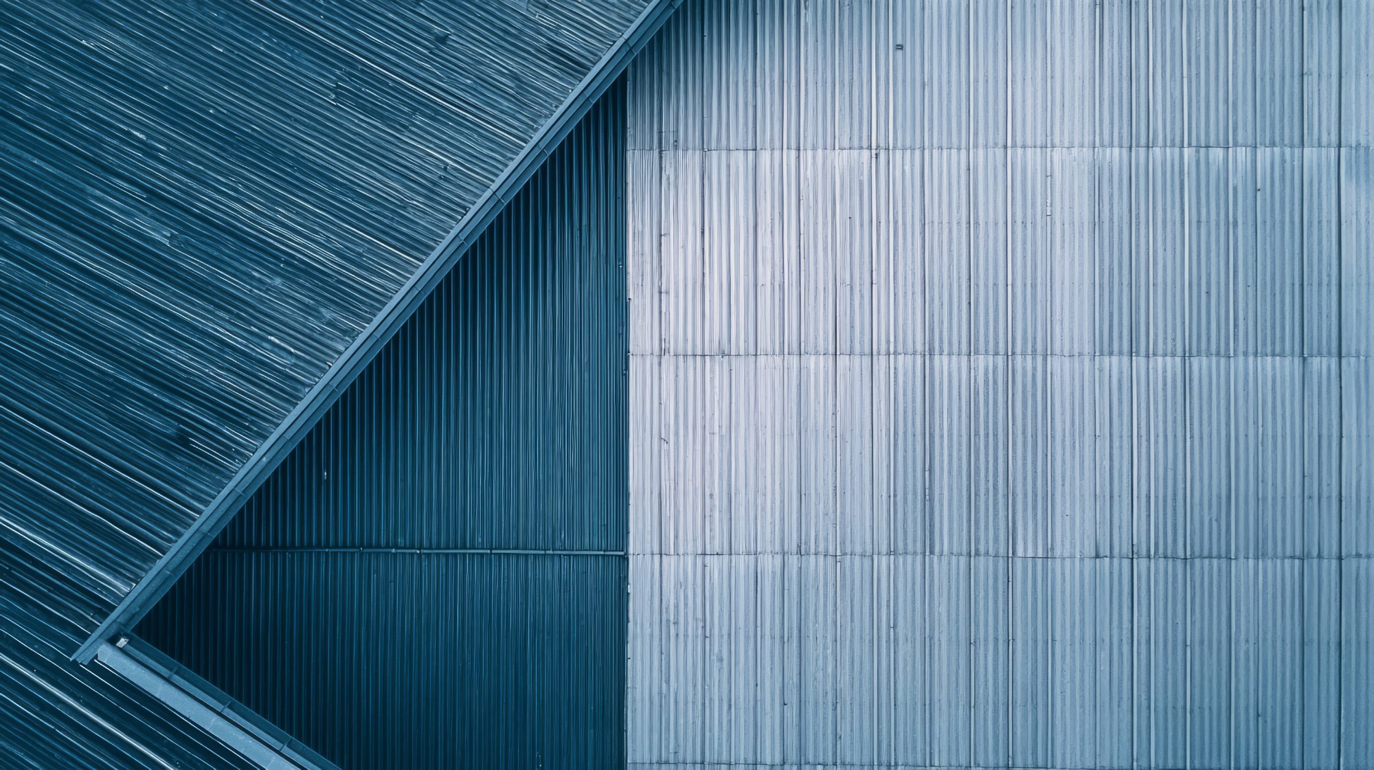 Aerial view of intersecting metal roofs in shades of blue and gray, forming geometric patterns. The image conveys a modern, industrial tone.
