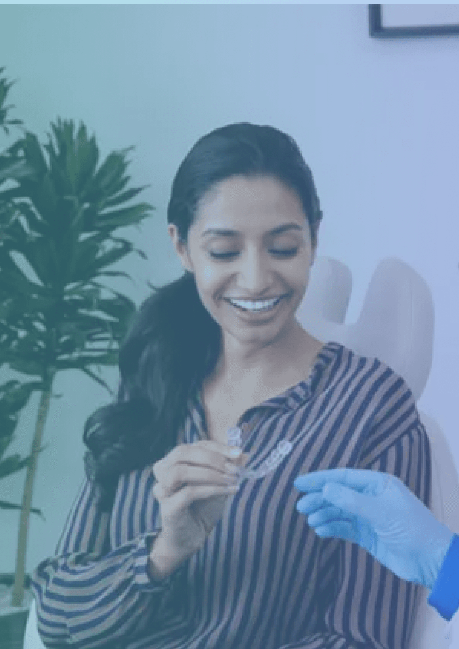 A smiling woman sitting in a dental chair, holding a clear aligner with a dentist's hand