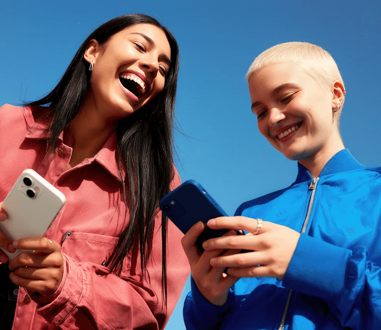 Two smiling people outdoors, one in a pink jacket holding a white phone, the other in blue with a buzz cut holding a blue phone, under a clear sky.