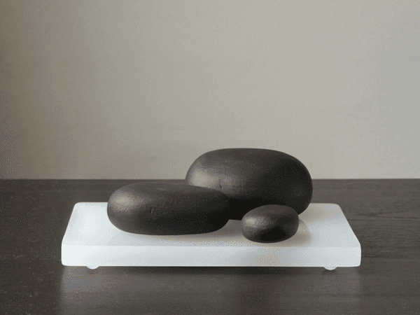 Three dark gray or black stones rest on a white rectangular base. The largest stone is positioned slightly behind the other two, which are closer together. The base sits on a dark surface against a light gray wall. The stones have a smooth, matte finish and cast subtle shadows on the surface.