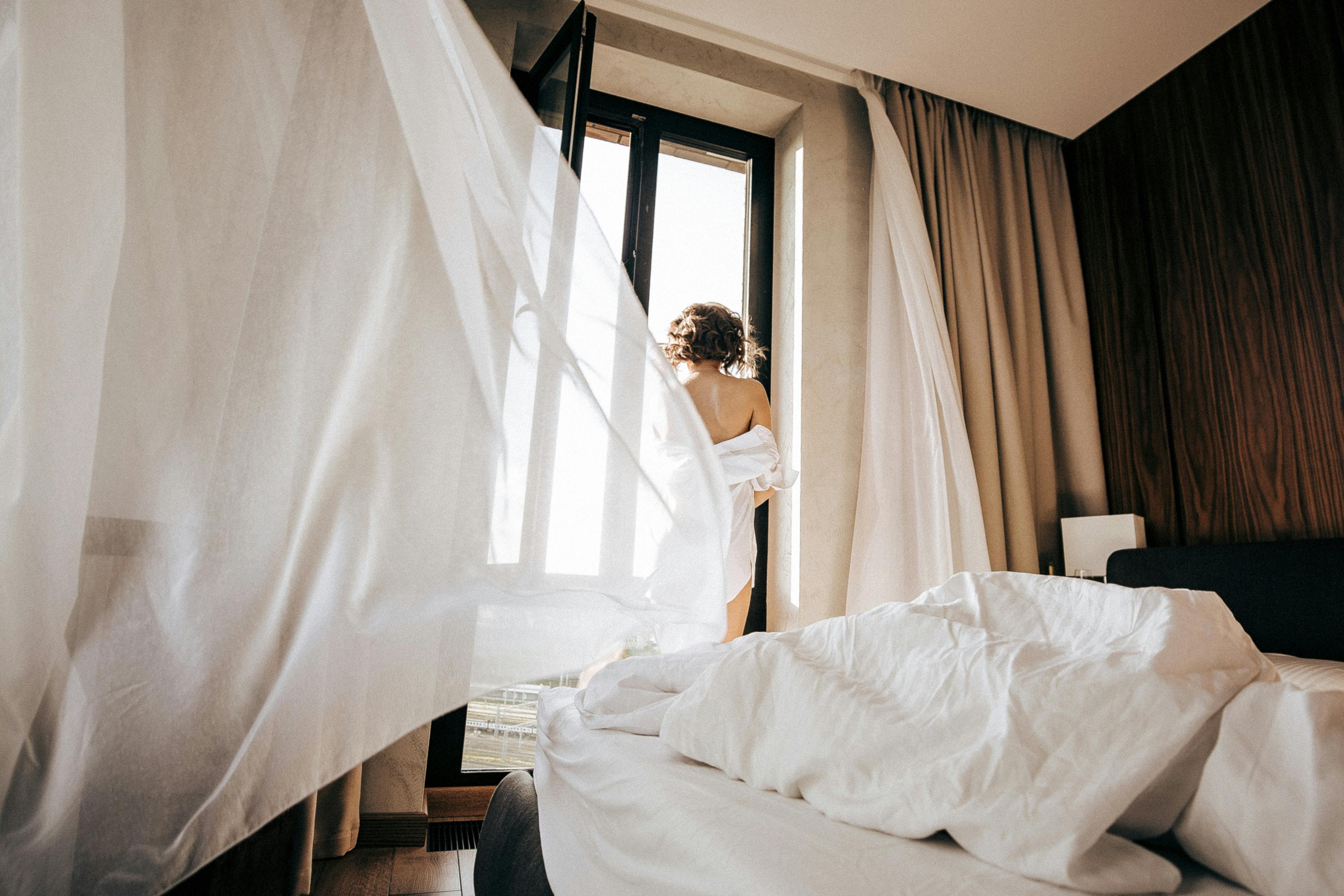 A woman stands by an open window and the wind blows the curtains