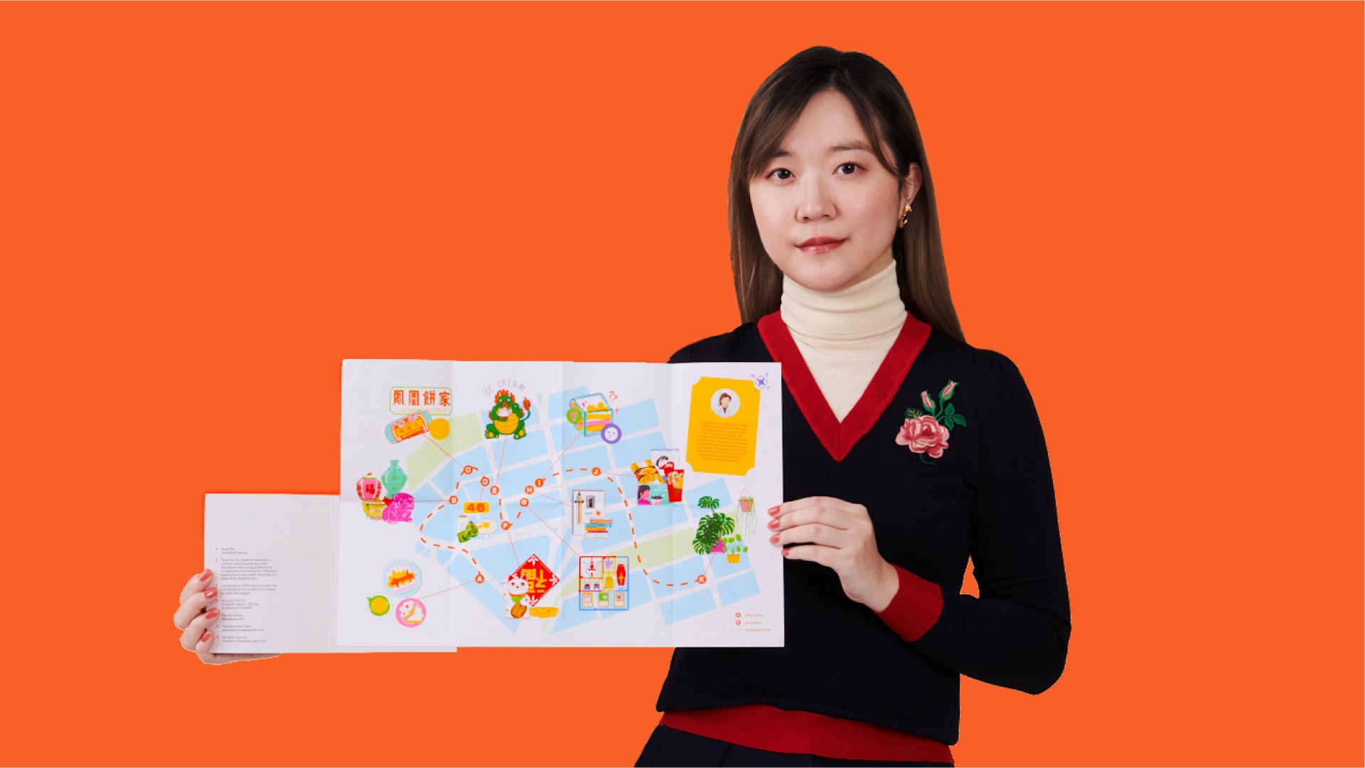 A woman with dark hair and a black sweater with a red trim at the neckline holds open a colorful illustrated map with Chinese characters and illustrations. She wears a cream-colored turtleneck underneath the sweater and a dark skirt. The background is solid orange.