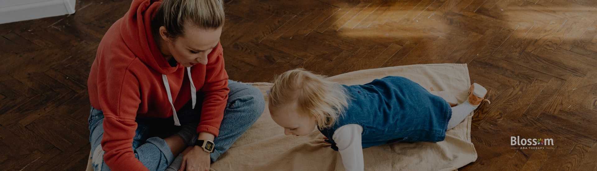 A woman in a red hoodie plays with a young child on a blanket-covered floor.