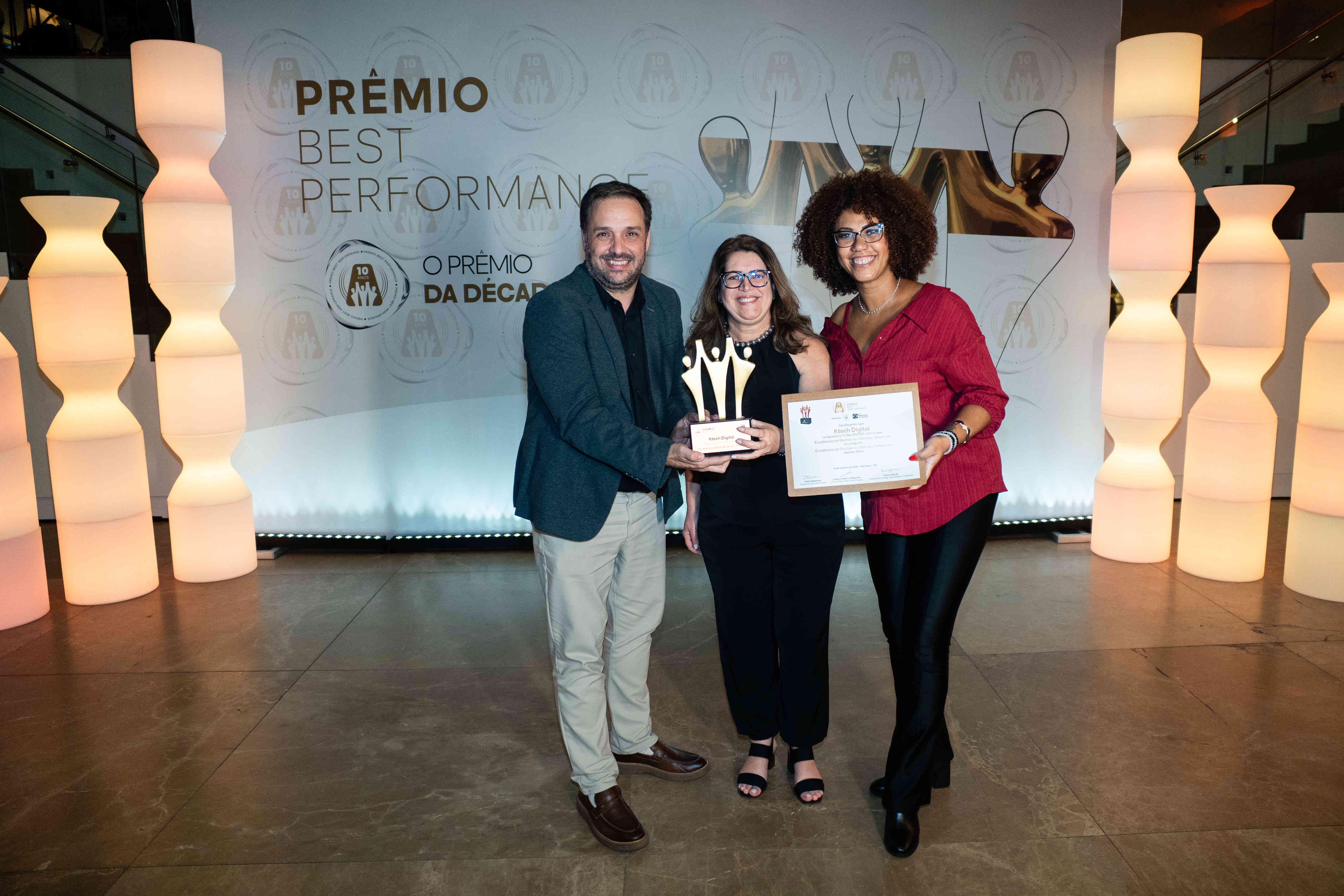 Executivos da Ktech Digital posando com troféu bronze e certificado no palco do Prêmio Best Performance 2025