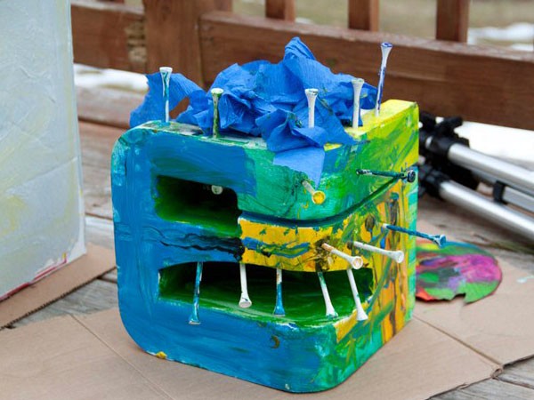 A square block of painted wood with blue, green, and yellow paint is decorated with blue tissue paper on top and white sticks protruding from its sides. The block rests on cardboard atop a wooden surface, with a paint palette visible to the right. A wooden fence and part of a tripod are in the background.