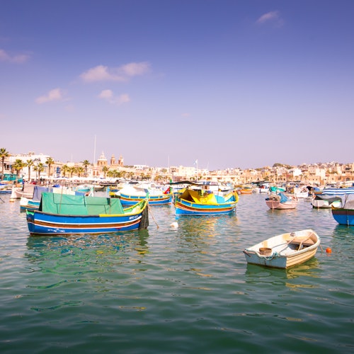 Гавань с красочными деревянными лодками, плавающими по спокойной воде под ясным, ярким небом; вдалеке видны здания городка.