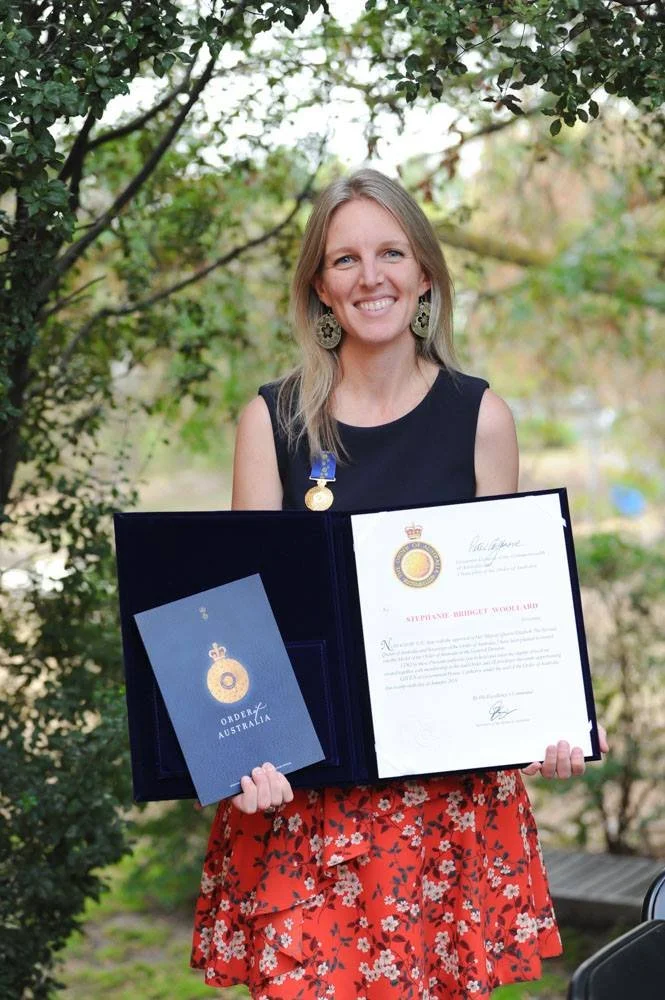 Person wearing a blue puffer jacket and red backpack standing in front of a light blue building on a city street.