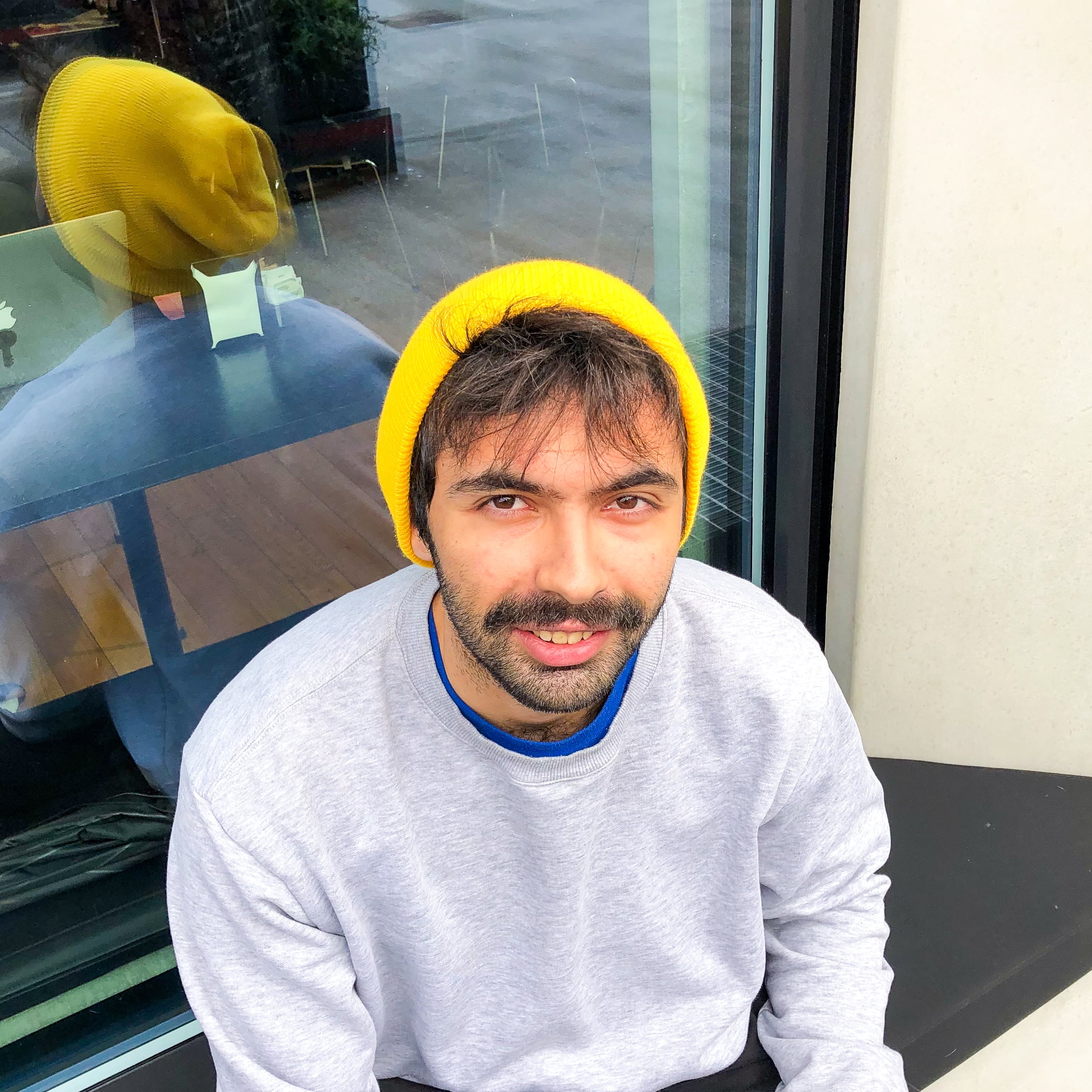 Een persoon met een gele muts en een grijze trui zit op een rand en glimlacht naar de camera.