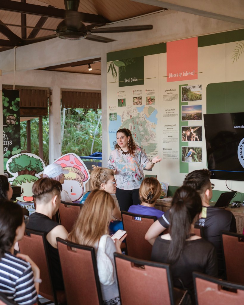 A woman presenting a trail guide in a resort setting, with guests attentively listening and colorful educational displays in the background.