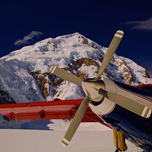 一架紅色機翼的螺旋槳飛機停在雪地上，背景是被雪覆蓋的山脈，在黑暗的天空下。