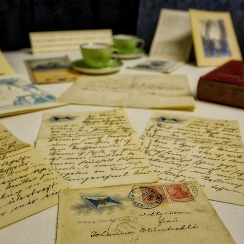 Vintage letters and postcards on a table, surrounded by green teacups, framed photos, and a red book.