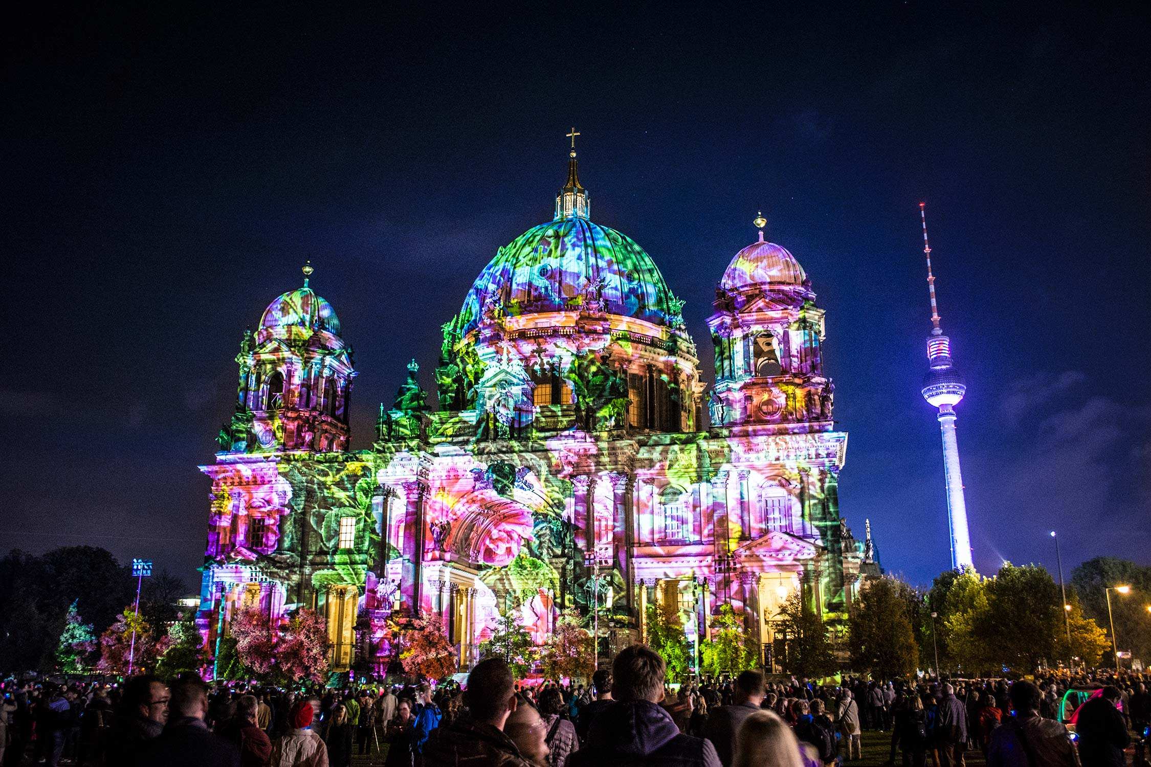 A large domed cathedral illuminated with colorful light projections and a crowd of people in front at night.