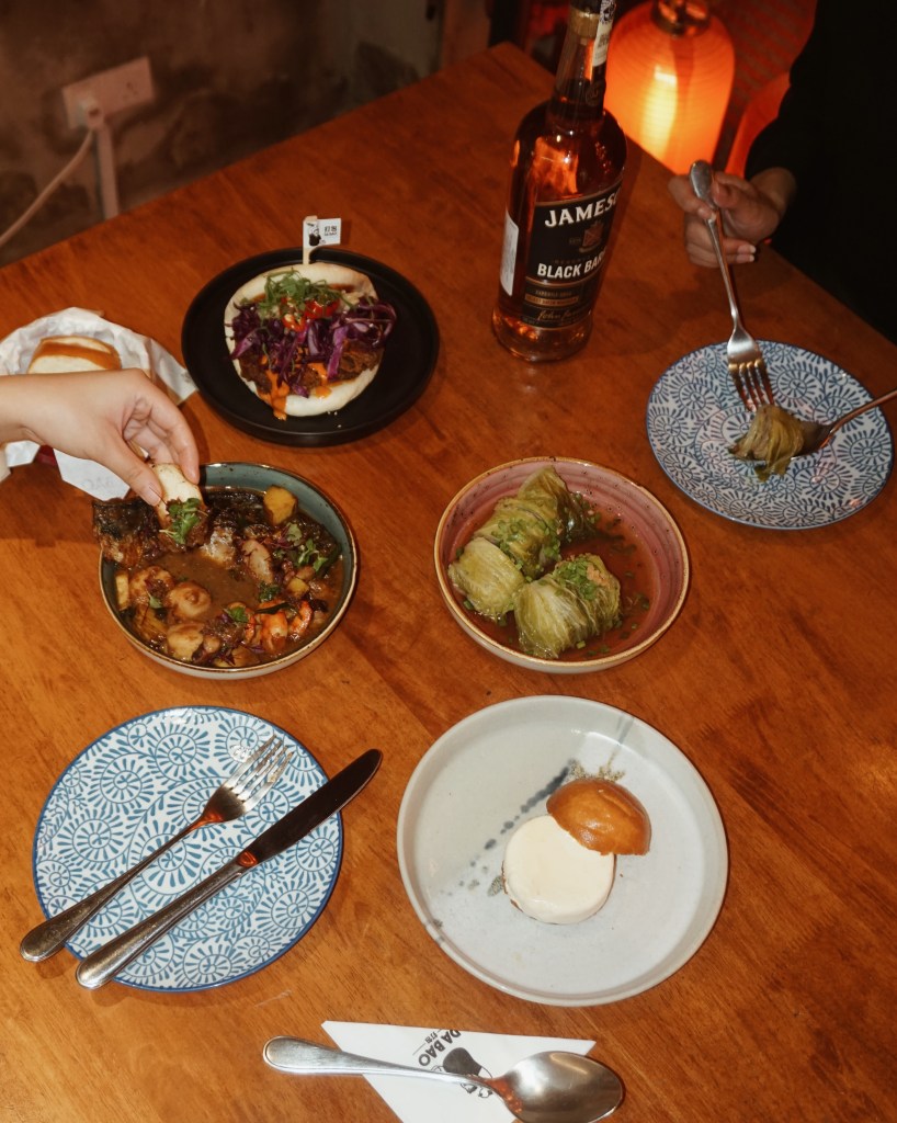 A table setting featuring various dishes and a bottle of Jameson Black Barrel whiskey. The dishes include a vibrant bao with vegetables, a bowl of seafood and meat stew, stuffed cabbage rolls, and a dessert with a bun and cream. Cutlery is neatly arranged on plates.