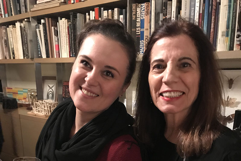 A photo of two smiling women sitting in front of a book shelf.