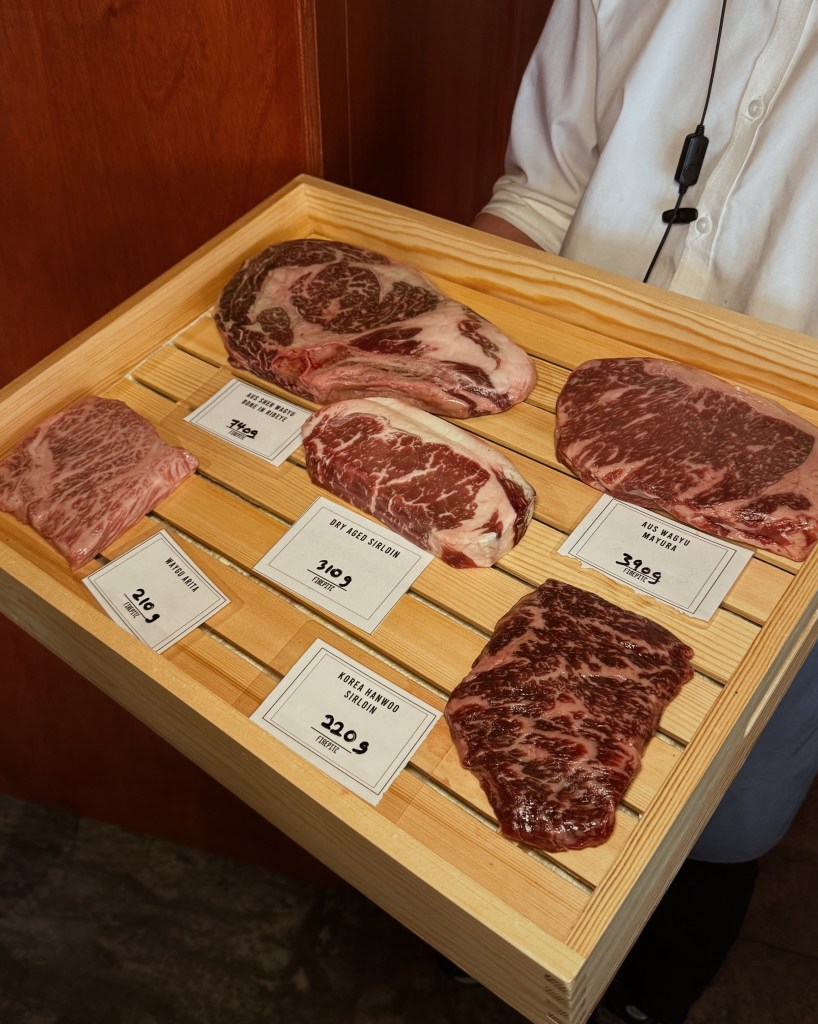 A selection of premium dry-aged steaks displayed on a wooden tray, with price labels for each cut. The cuts include Kobe beef, Hanwoo sirloin, and Australian Wagyu.
