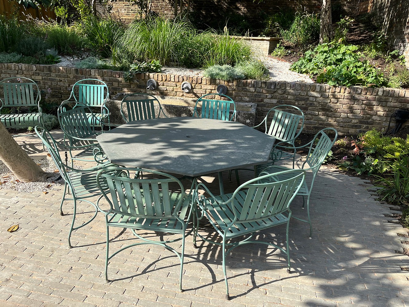 A set of green metal patio furniture arranged on a stone surface, surrounded by greenery and plants.