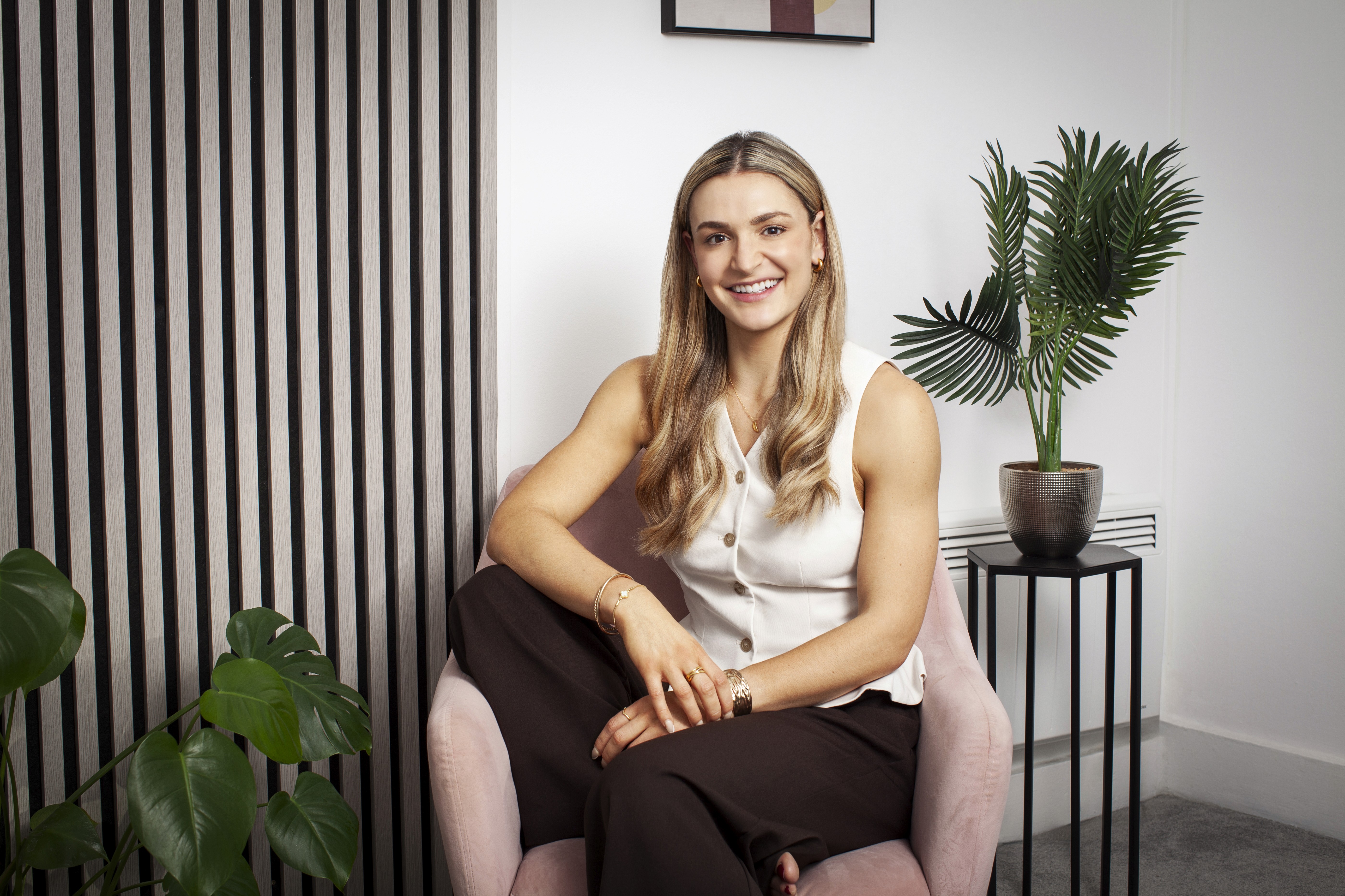 Woman sitting in a chair, smiling at the camera, with indoor plants and a modern, minimalist background.