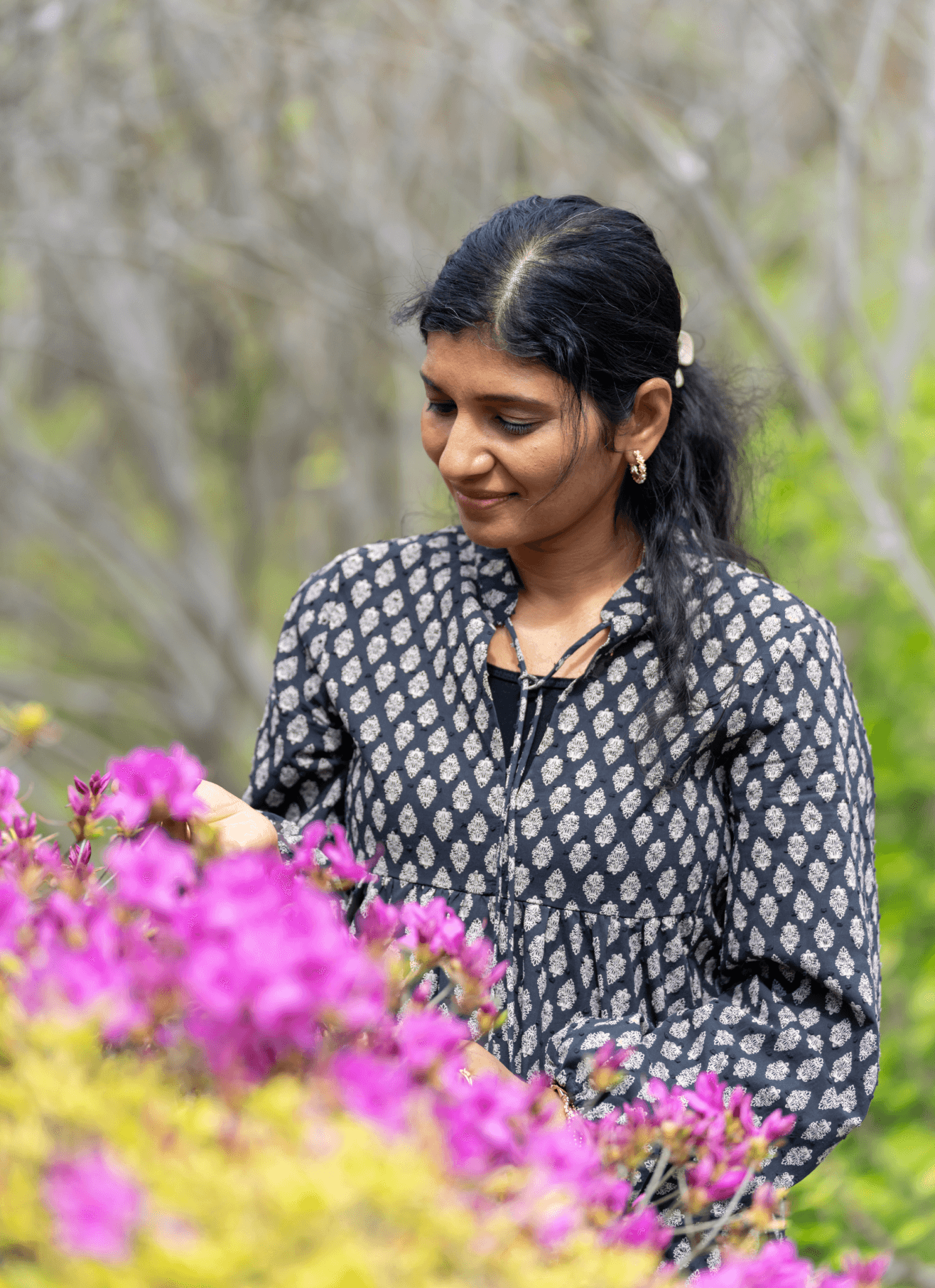 Sunitha Vinod in a patterned dress admires vibrant pink flowers in a lush garden, embodying tranquility and the essence of nature, reminiscent of Ayurvedic principles; associated with Ayurvedic consultation Louisville.