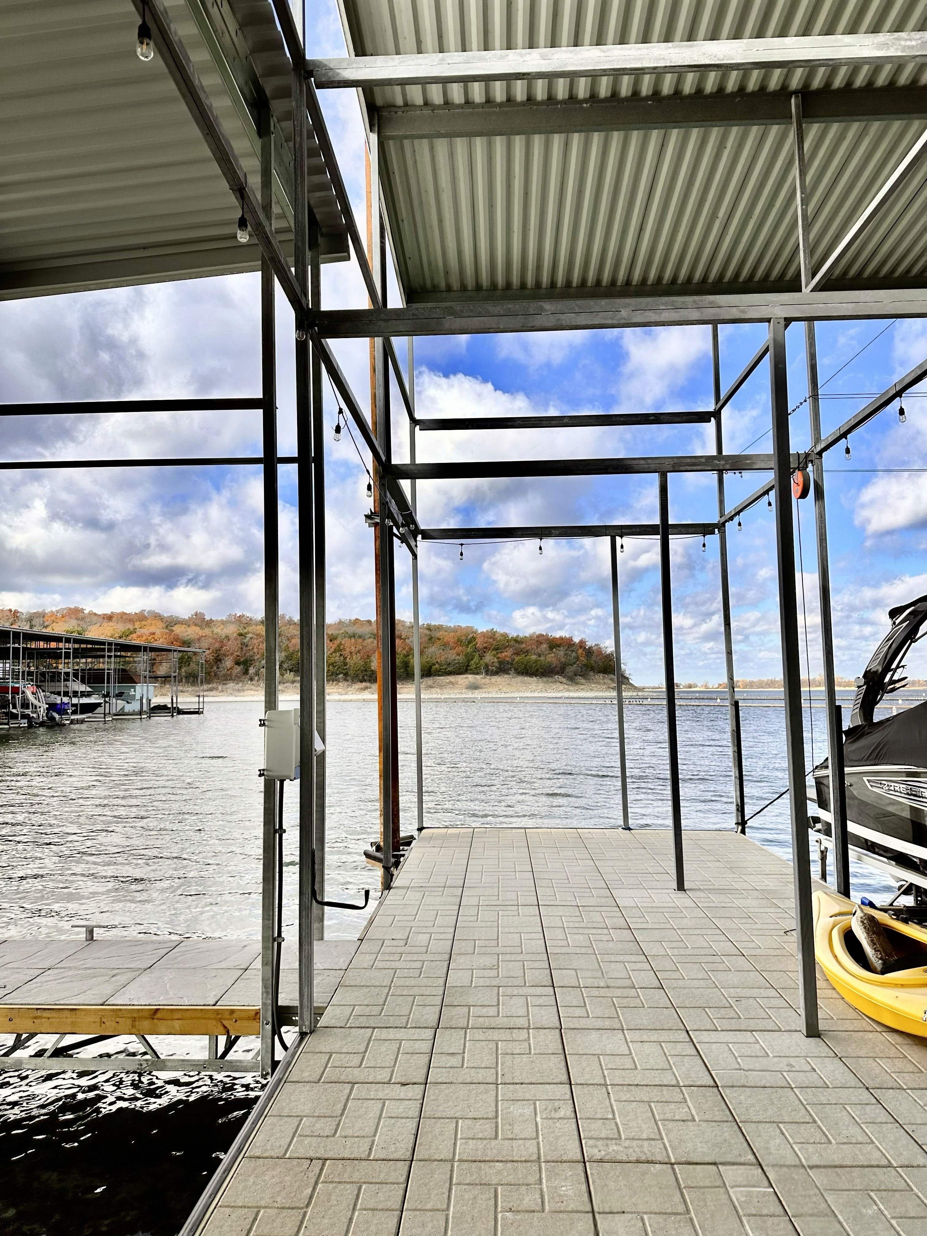 A waterside view of a boat dock with a tiled walkway extending toward the water, framed by a metal structure, and surrounded by a serene lake under a partly cloudy sky.