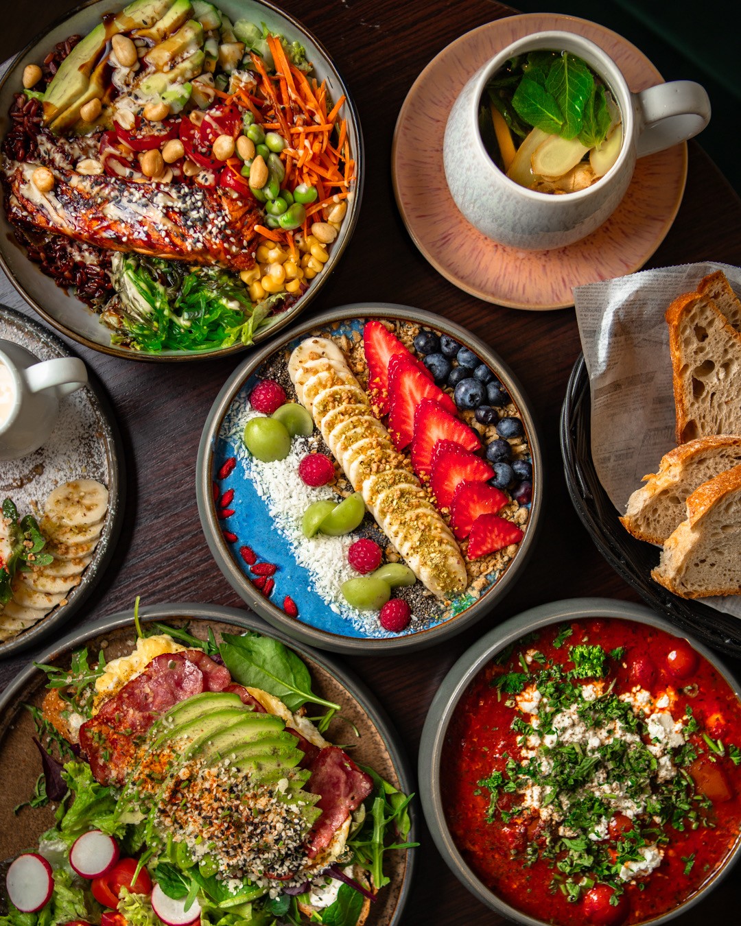 A sandwich wrapped in paper, served with chips and a drink, on a wooden table surrounded by greenery.