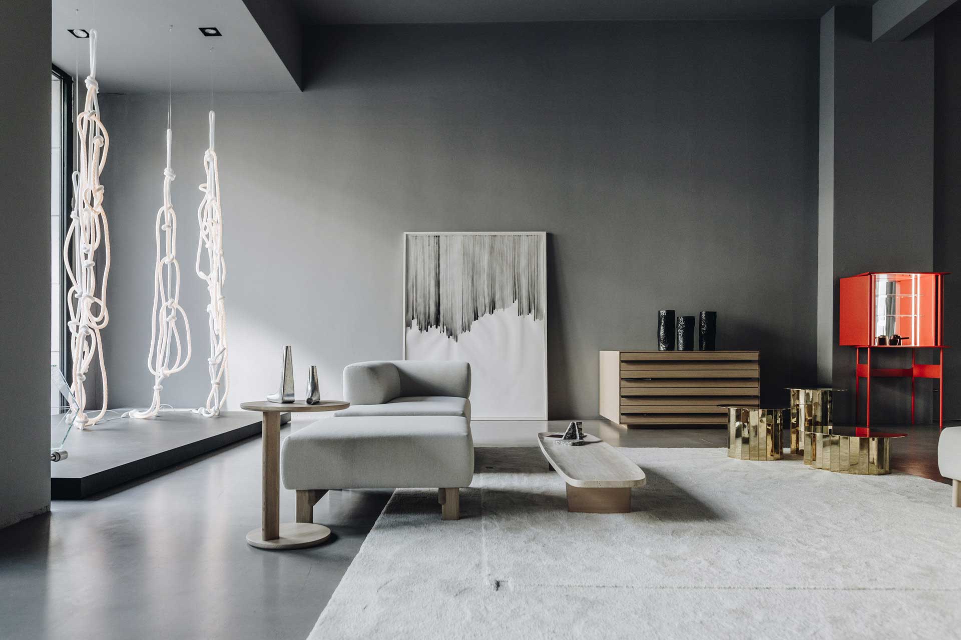 A large, modern room with gray walls and concrete floor. The space is furnished with a white sofa, a coffee table, a red cabinet, and two large, illuminated sculptural light fixtures made of rope hanging from the ceiling.
