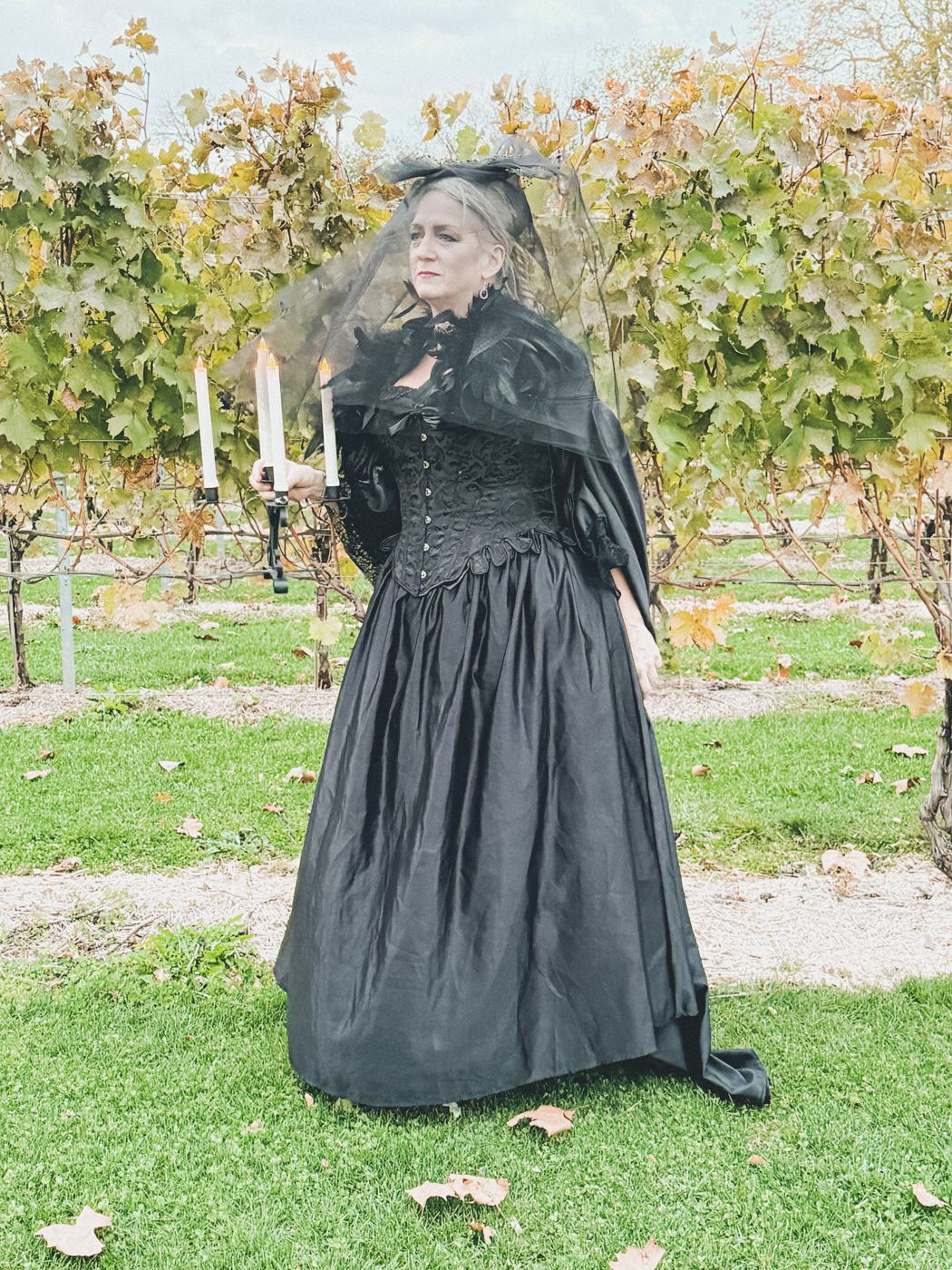 A guest in a black Victorian gown and veil holding a lit candelabra in the vineyard