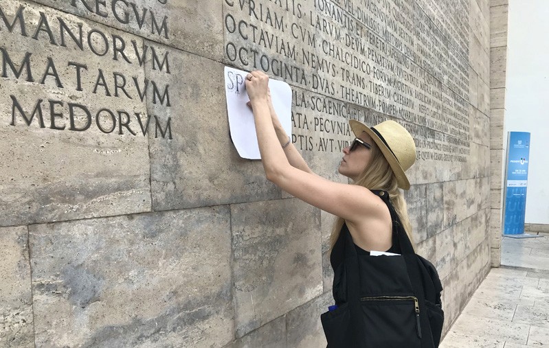 A girl copying on a piece of paper the letters engraved on a stone wall.