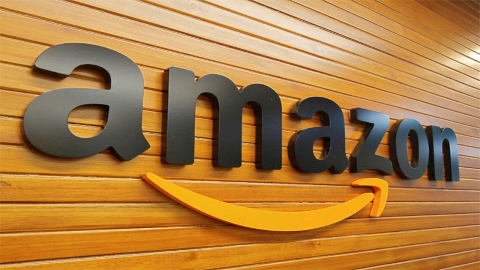 Amazon's logo, featuring bold black lowercase letters with a yellow arrow underneath, is displayed on a wooden wall background.