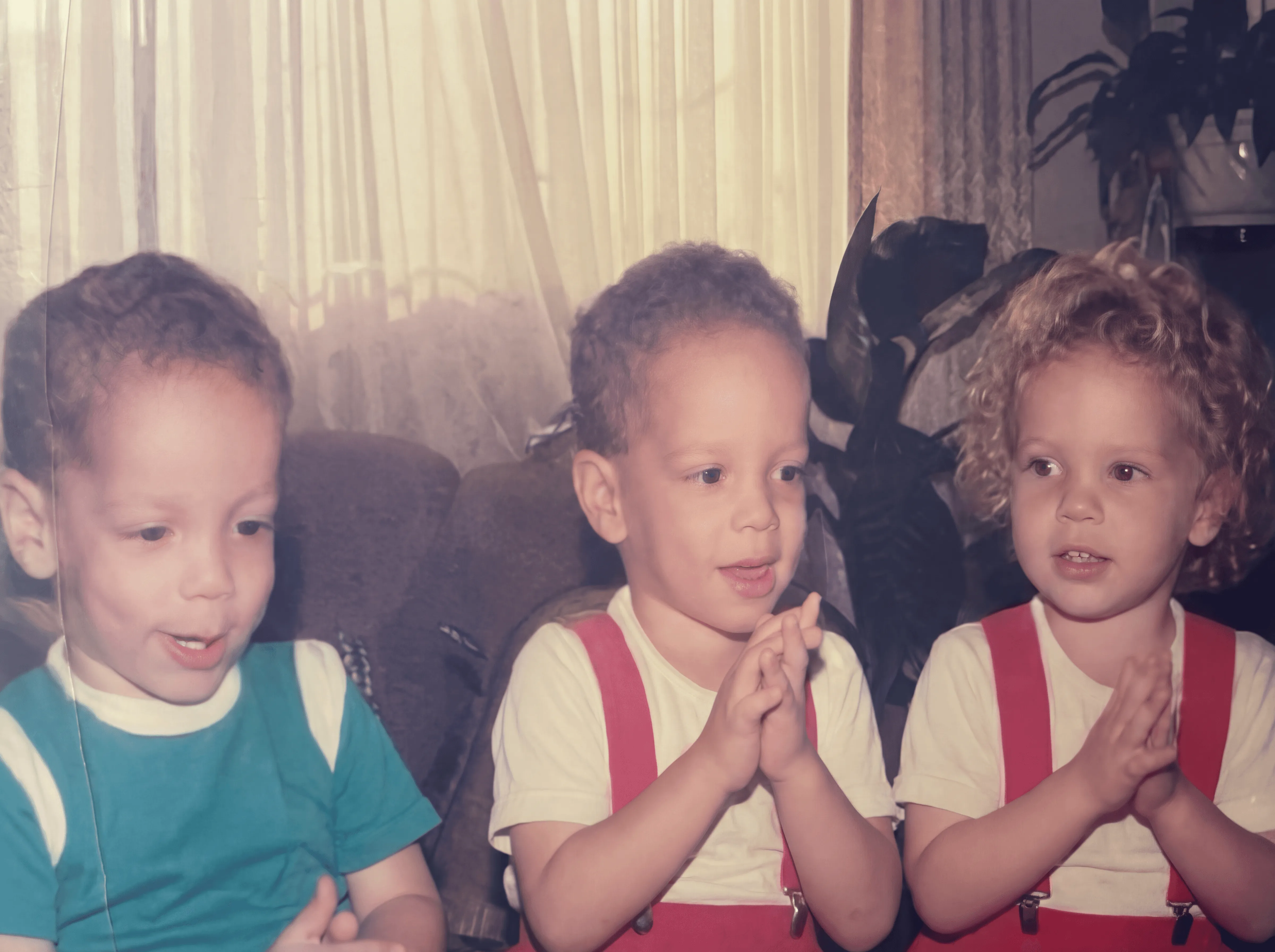 Tres niños pequeños sentados juntos en una sala, con las manos juntas como si aplaudieran o rezaran, frente a una ventana con cortinas claras y un ambiente familiar.