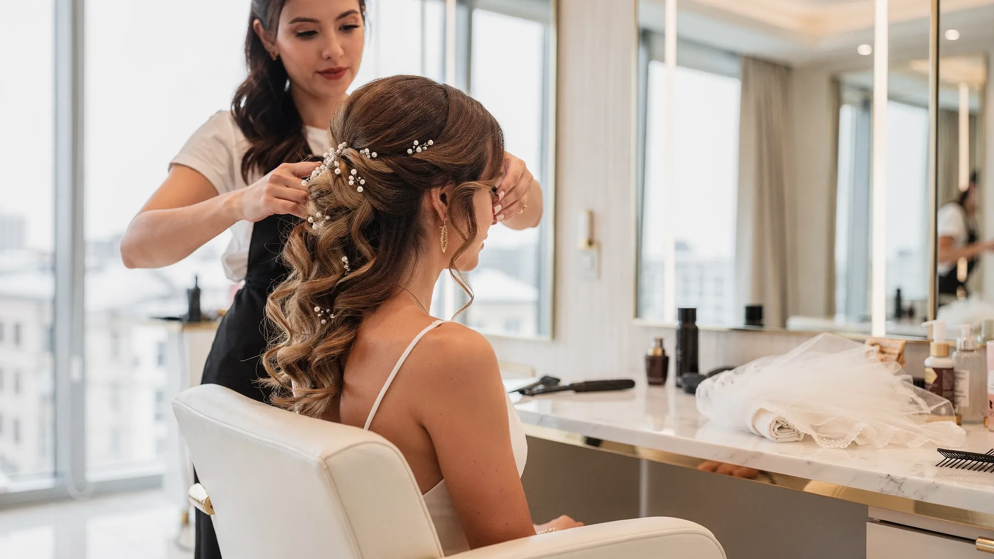 A bride seated in a salon chair with soft curled bridal hair, pearl hair pins, a veil nearby, and a stylist arranging the final face-framing pieces in a bright luxury salon.