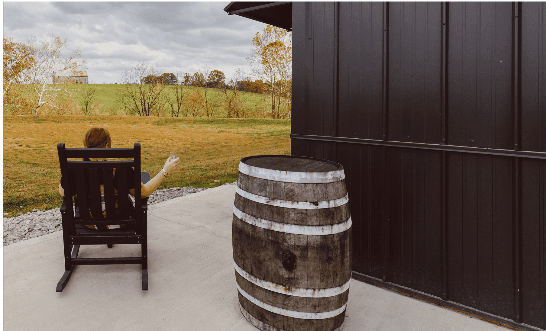 A woman sitting down facing away from the camera enjoying a drink and looking out at the fields.