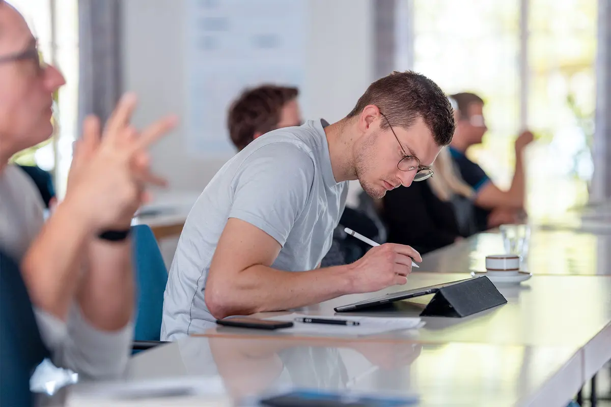 Ein junger Teilnehmer des SOP Notaufnahme Seminars mit Brille schreibt in einem hellen Klassenzimmer bei Tageslicht konzentriert in ein Notizbuch, umgeben von anderen Teilnehmenden, die dem Vortrag beiwohnen.