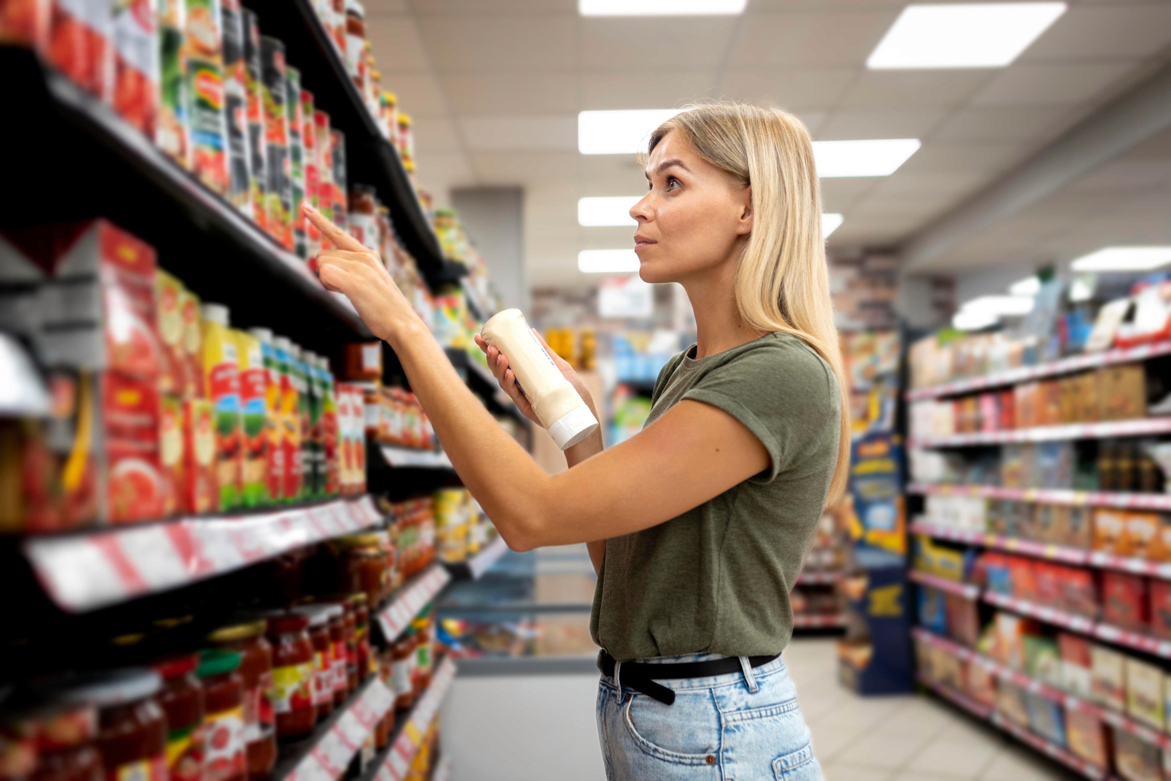 Una mujer en un pasillo de supermercado sostiene un producto en una mano mientras se estira para examinar algo en la estantería. Ella está mirando el establecimiento con atención, probablemente tomando una decisión de compra.