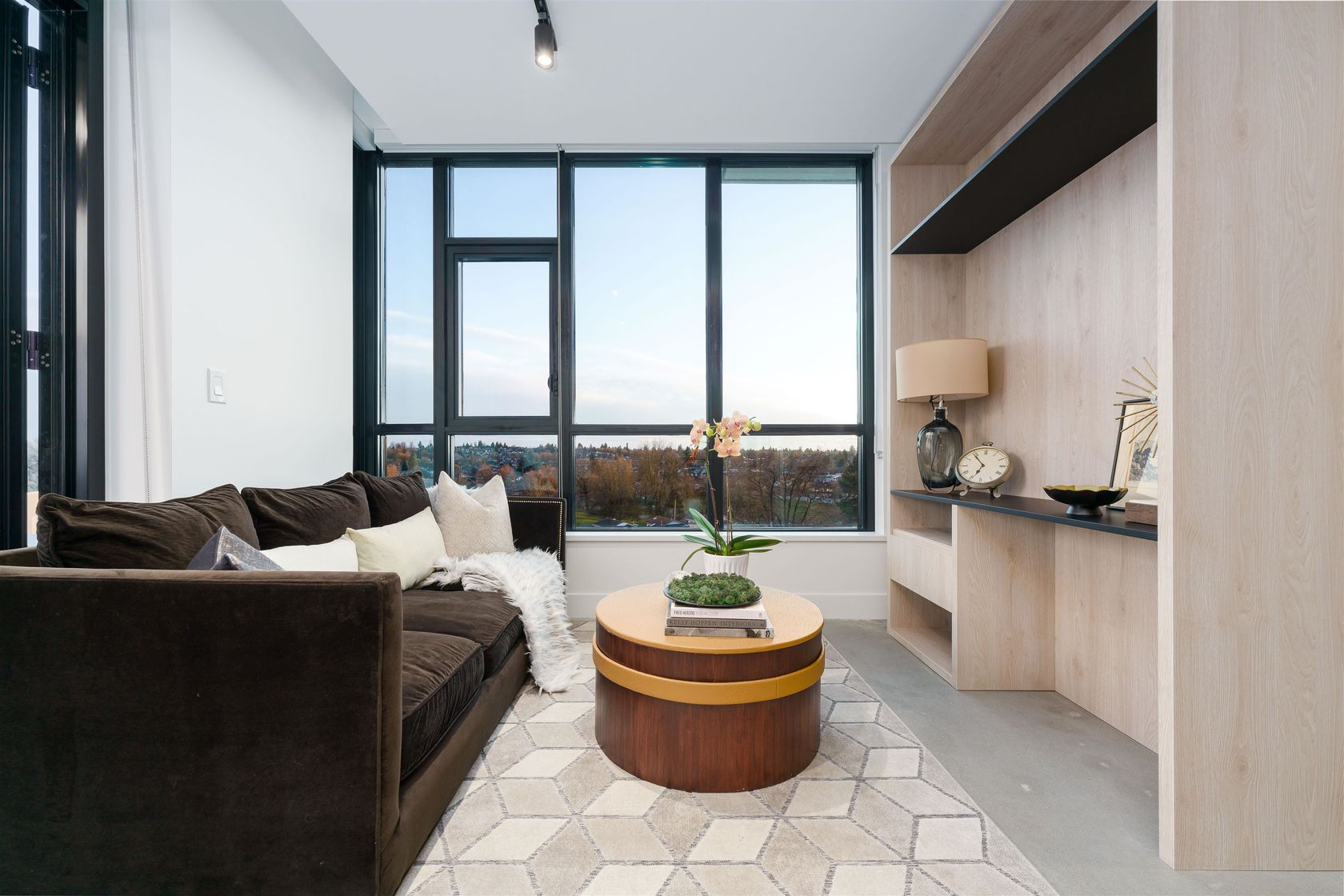 Modern living room featuring large windows, a couch, coffee table, and a sleek kitchen area. Bright and inviting space.